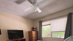 Ein Schlafzimmer mit einem Bett, einem Deckenventilator, einem Fenster mit Vorhängen, einem Fernseher auf einem Holzständer und einer kleinen Kommode.
