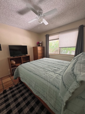 Een slaapkamer met een bed, een plafondventilator, een raam met gordijnen, een televisie op een houten standaard en een klein kastje.