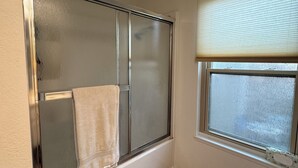 Ein Badezimmer mit Duschkabine aus Glas, einem Handtuch, das an der Tür hängt, einem Fenster mit Jalousien und einem gemusterten Teppich auf dem Boden.