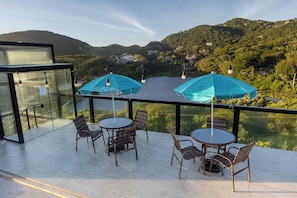 A terrace with outdoor seating, a glass railing, and a view of a lake and hills. - Casa Oceano - Praia do Rosa (Imbituba)