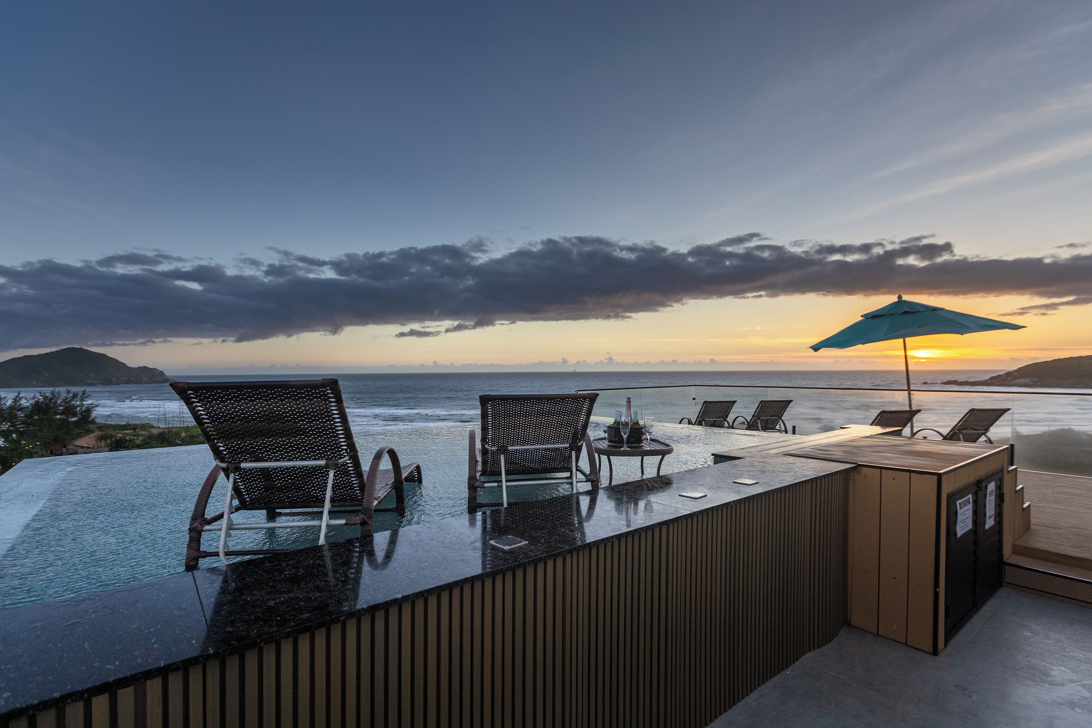 Piscina infinita com cadeiras e mesa, com vista para o oceano e um pôr do sol.