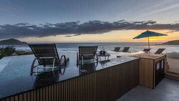 Piscina infinita com cadeiras e mesa, com vista para o oceano e um pôr do sol.