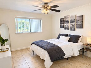 Une chambre à coucher comprenant un lit, un ventilateur de plafond, une fenêtre avec des stores, une table de chevet avec une lampe et des œuvres d’art accrochées au mur.