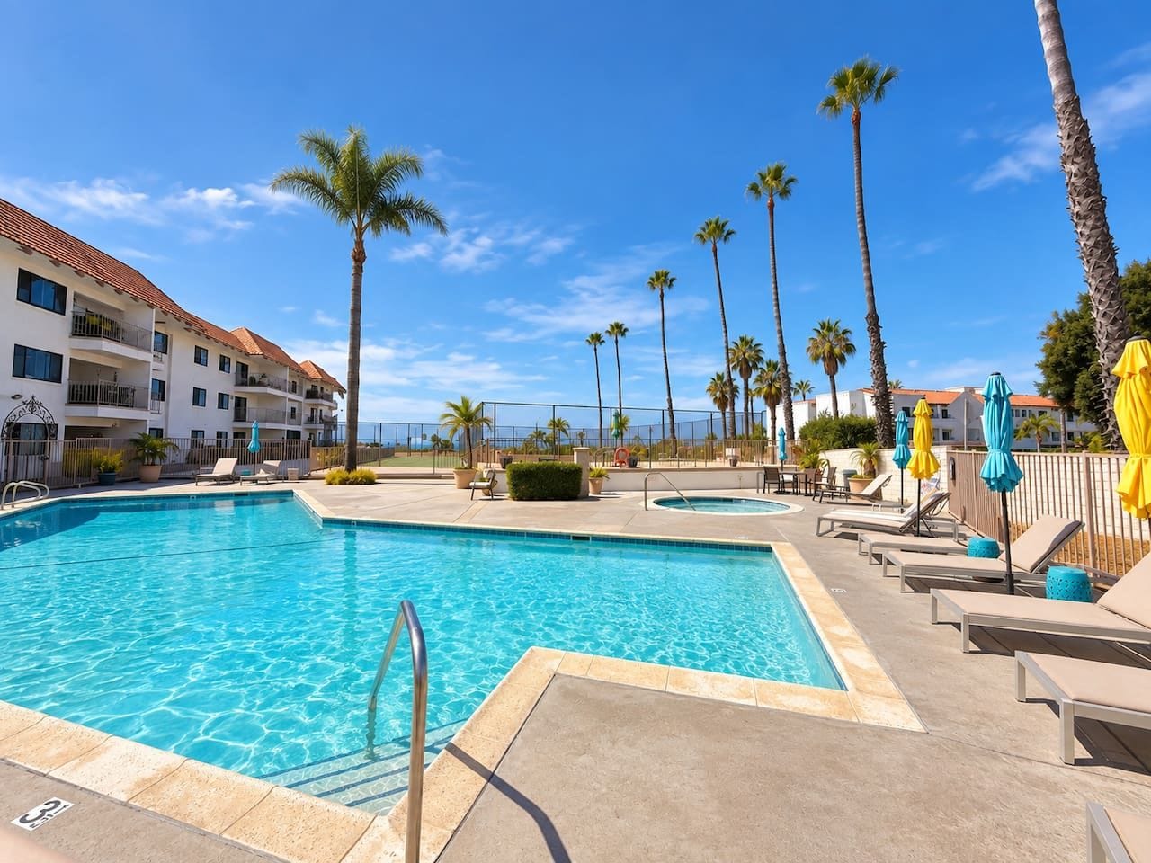 A swimming pool with lounge chairs and umbrellas, surrounded by palm trees and a building with balconies.