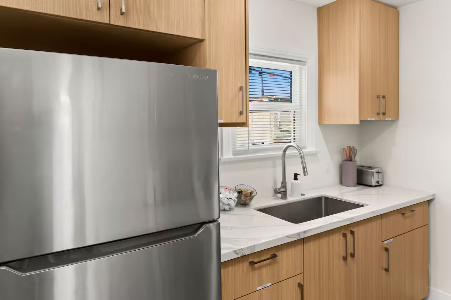 A kitchen with a stainless steel refrigerator, wooden cabinets, a sink, and a window with blinds.