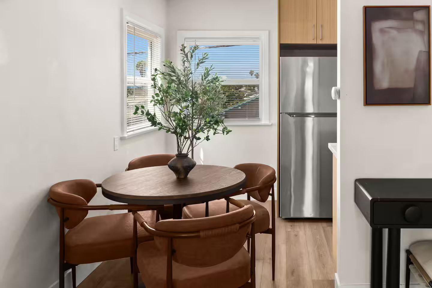 Una cocina compacta con refrigerador de acero inoxidable, una mesa de comedor con cuatro sillas y una planta en el alféizar.