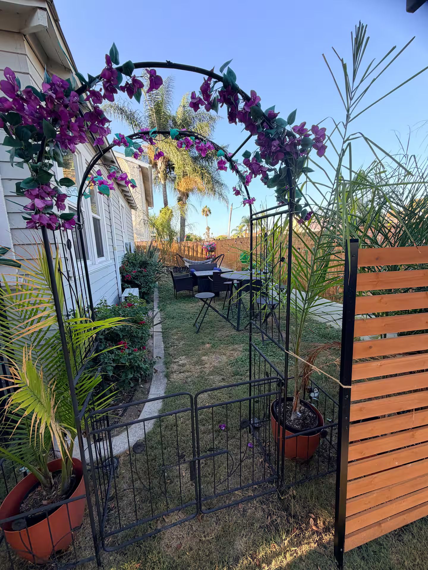 Un jardin agrémenté d’une pergola recouverte de fleurs violettes, d’une clôture en bois et d’une table avec des chaises.