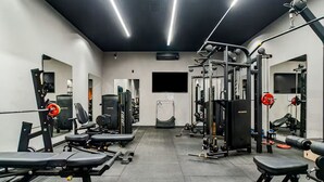 Academia com diversos equipamentos de exercício, espelhos e uma tela de televisão.