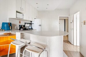 A modern kitchen with white cabinetry, a curved island, and two wicker bar stools. - Apartment with heated pool and gym in Alphaville - Barueri - São Paulo (Barueri)