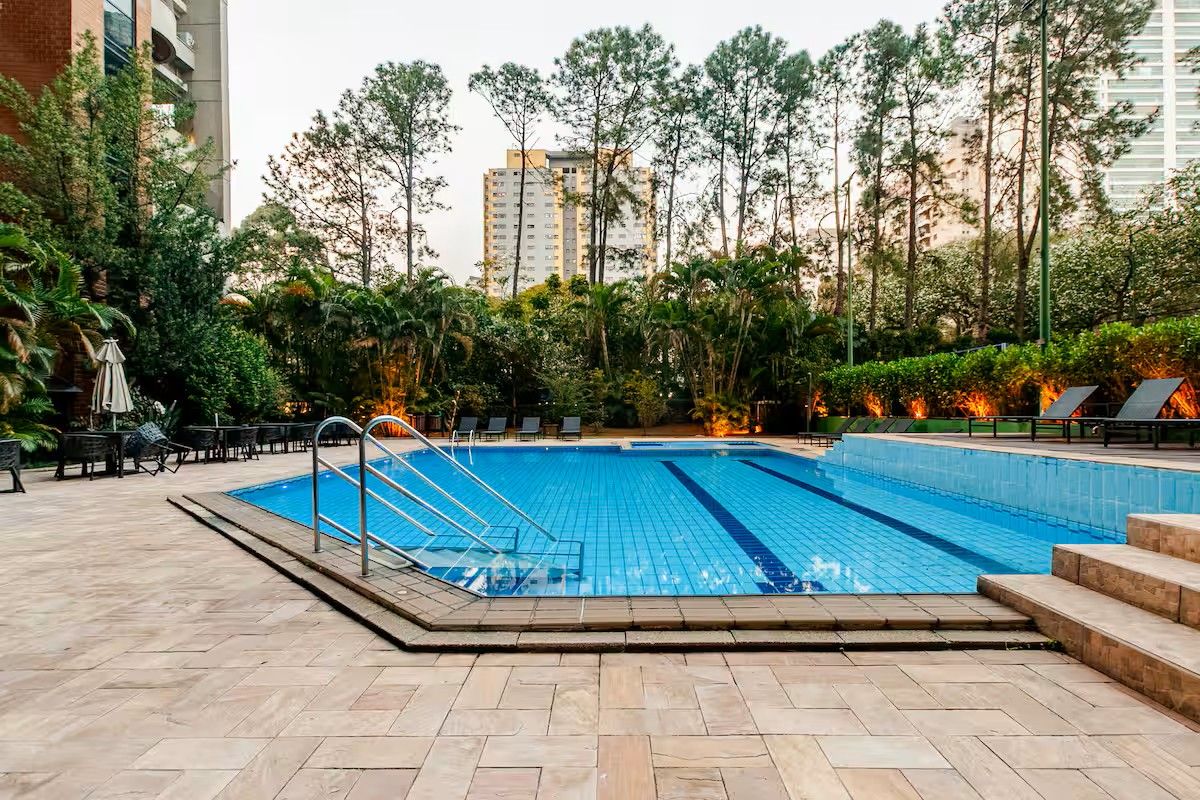 A swimming pool with a tiled deck, surrounded by greenery and tall buildings in the background.