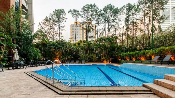 Piscina com deck ladrilhado, cercada por vegetação e prédios altos ao fundo.