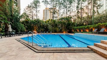 A swimming pool with a tiled deck, surrounded by greenery and tall buildings in the background.