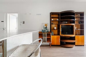 A modern living room with a curved wooden entertainment unit, a flat-screen TV, and a wicker chair. - Apartment with heated pool and gym in Alphaville - Barueri - São Paulo (Barueri)
