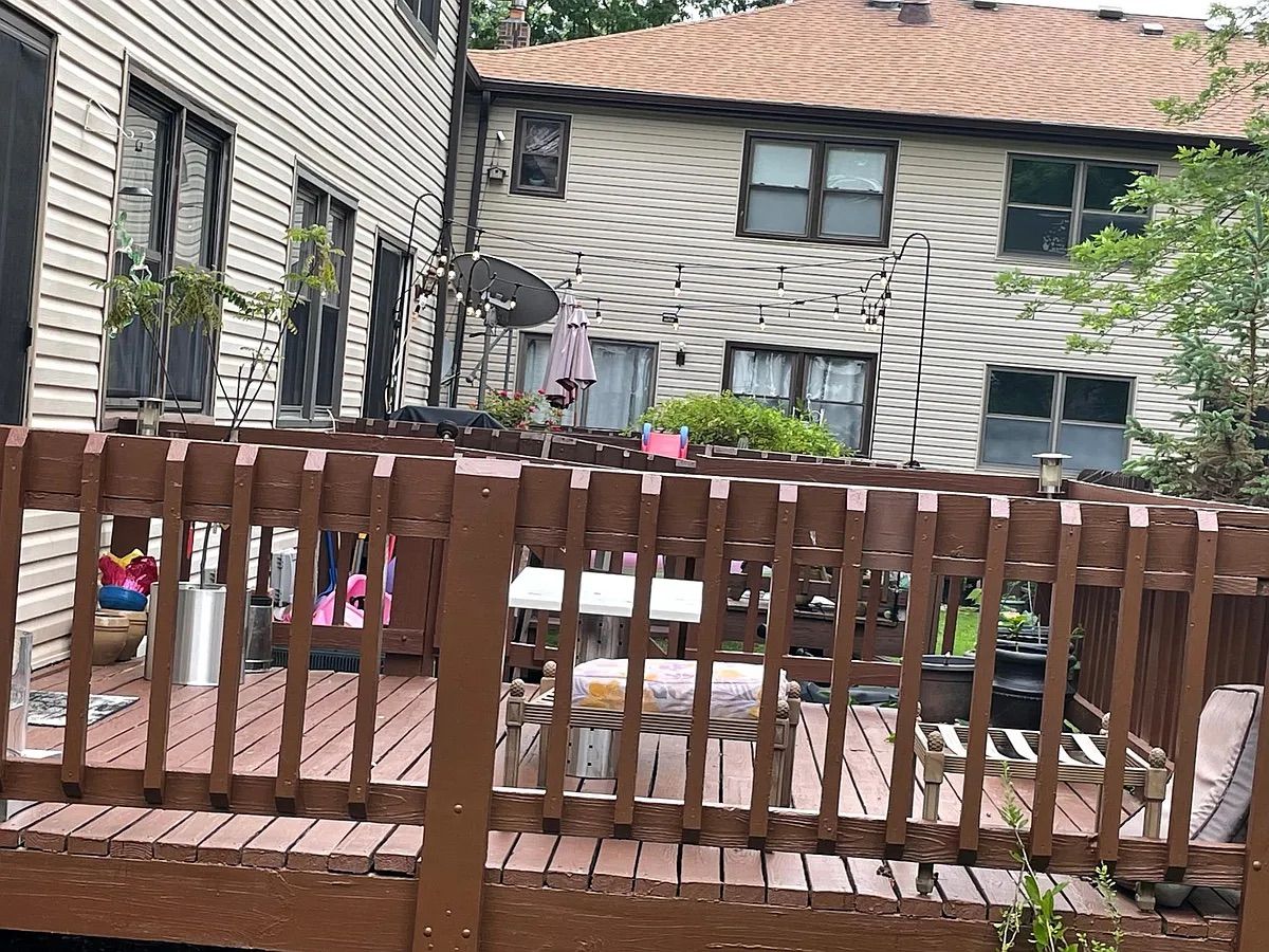 Um deck de madeira com um banco, cercado por uma grade, e casas ao fundo.