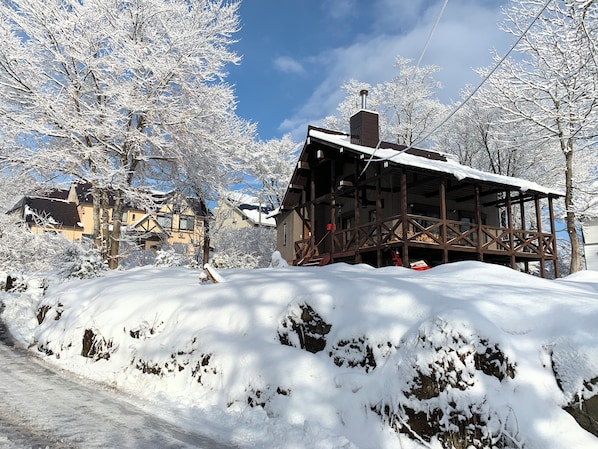 A wooden cabin with a snow-covered roof, surrounded by snow-laden trees and a clear sky.