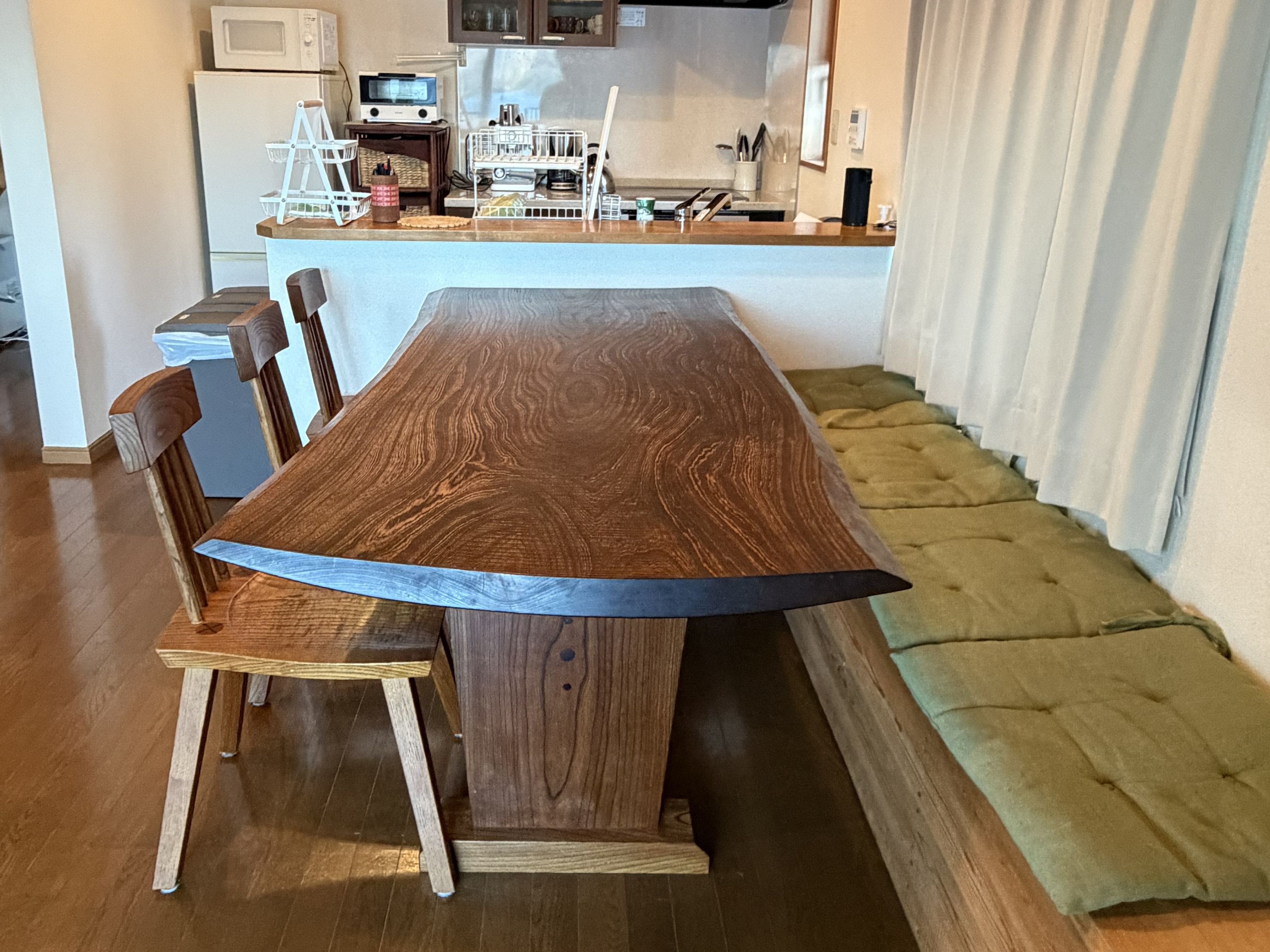 A wooden dining table with chairs, a green cushioned bench, and a kitchen area in the background.