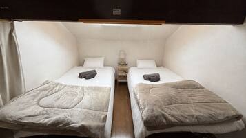 A compact bedroom with two beds, a bedside table with a lamp, and a small shelf.