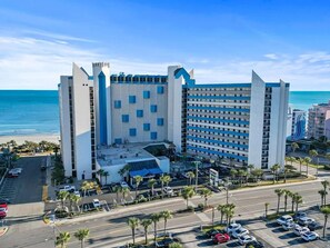 A hotel with a modern design, featuring a prominent blue and white color scheme, located near a beach with a parking lot and palm trees. - Family-Friendly 3BR | Indoor Pools & Arcade (Myrtle Beach)