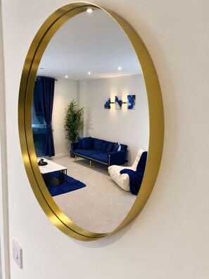 Ein Wohnzimmer mit einem blauen Sofa, einem weißen Sessel, einem kleinen Couchtisch und einem großen Spiegel mit goldener Rahmung.