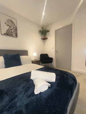 A neatly made bed with a dark blue cover, a leopard print artwork on the wall, a potted plant on a shelf, a black chair, and a closed door. - Manchester City Centre, Spacious, Luxury, Apartment (Manchester)