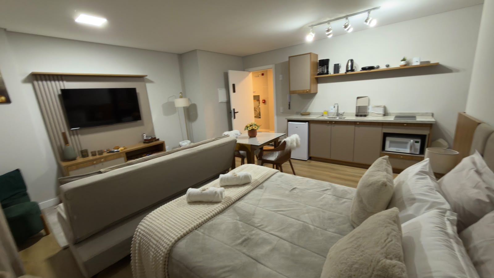 A modern hotel room with a bed, a TV, a kitchenette, and a dining area.