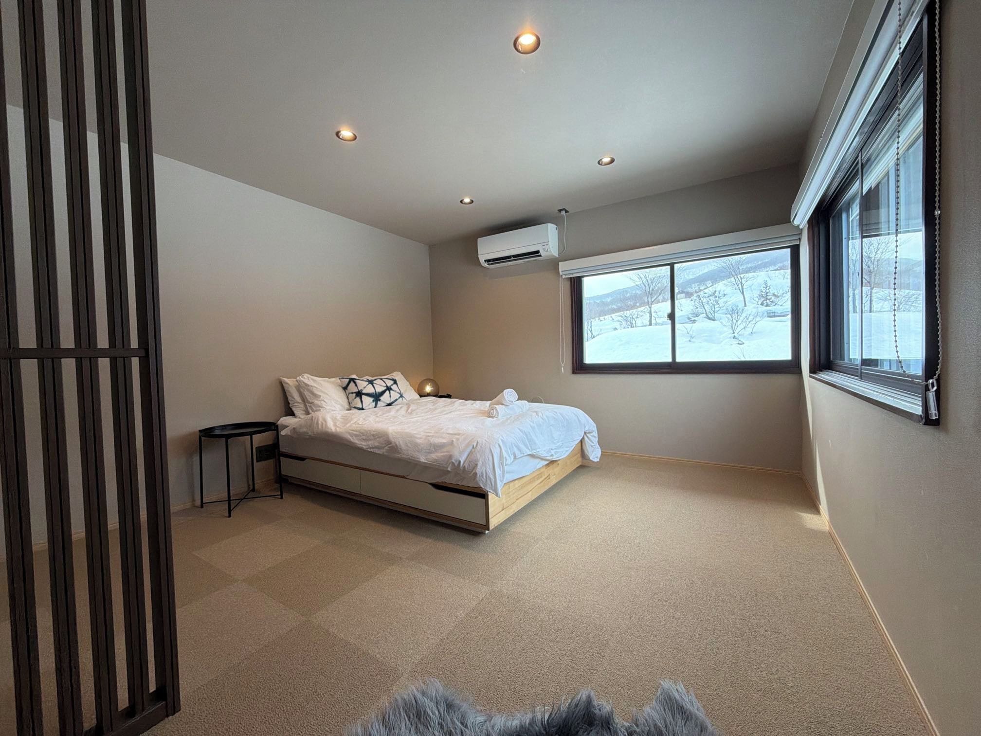 A bedroom with a bed, a small table, an air conditioning unit, and a window with a view of a snowy landscape.