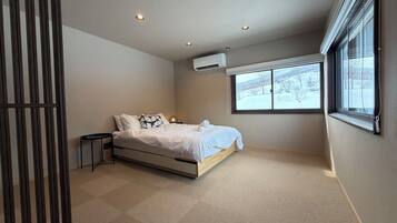 A bedroom with a bed, a small table, an air conditioning unit, and a window with a view of a snowy landscape.