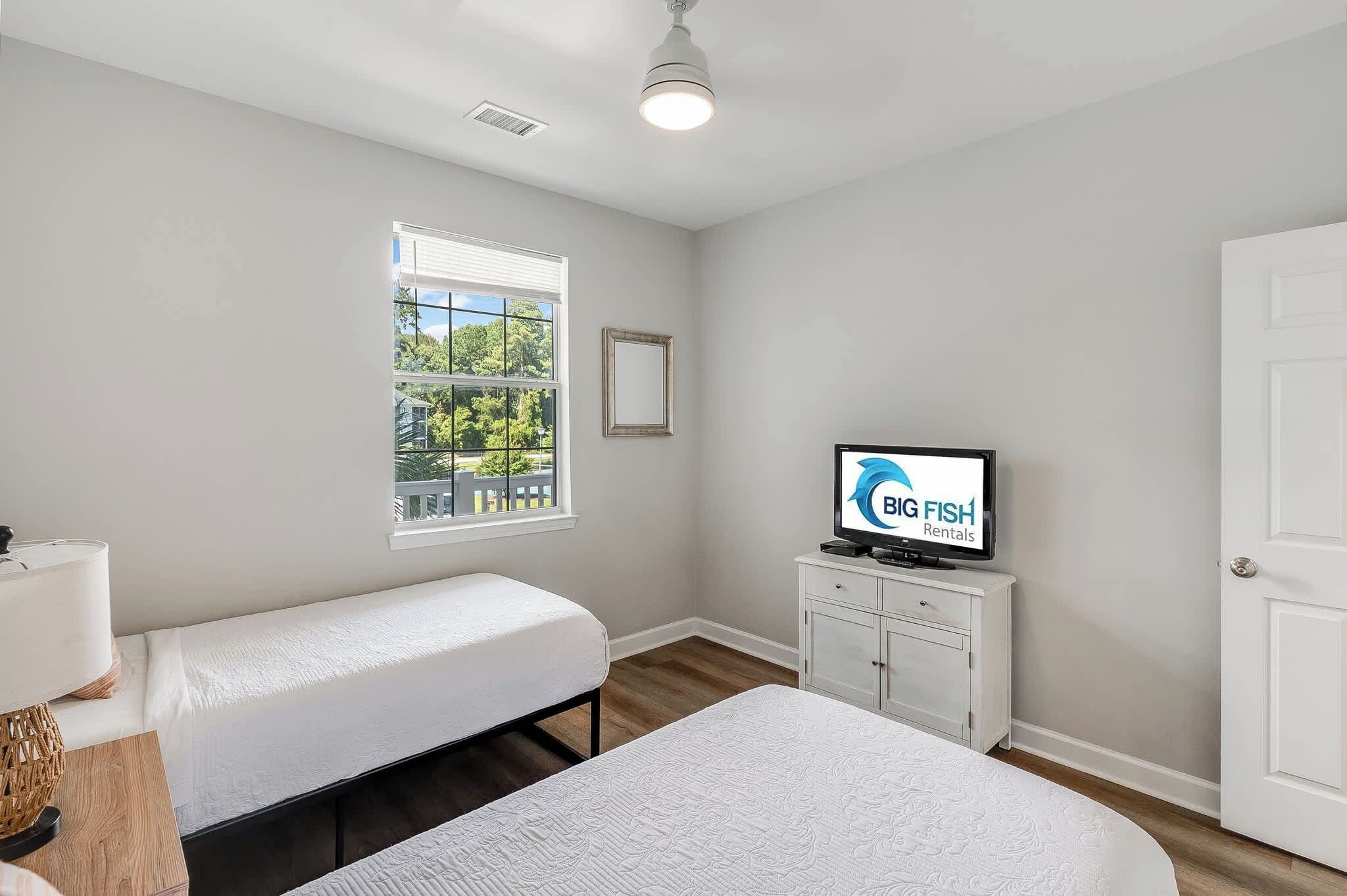 A bedroom with two single beds, a television on a cabinet, and a window with a view of greenery.