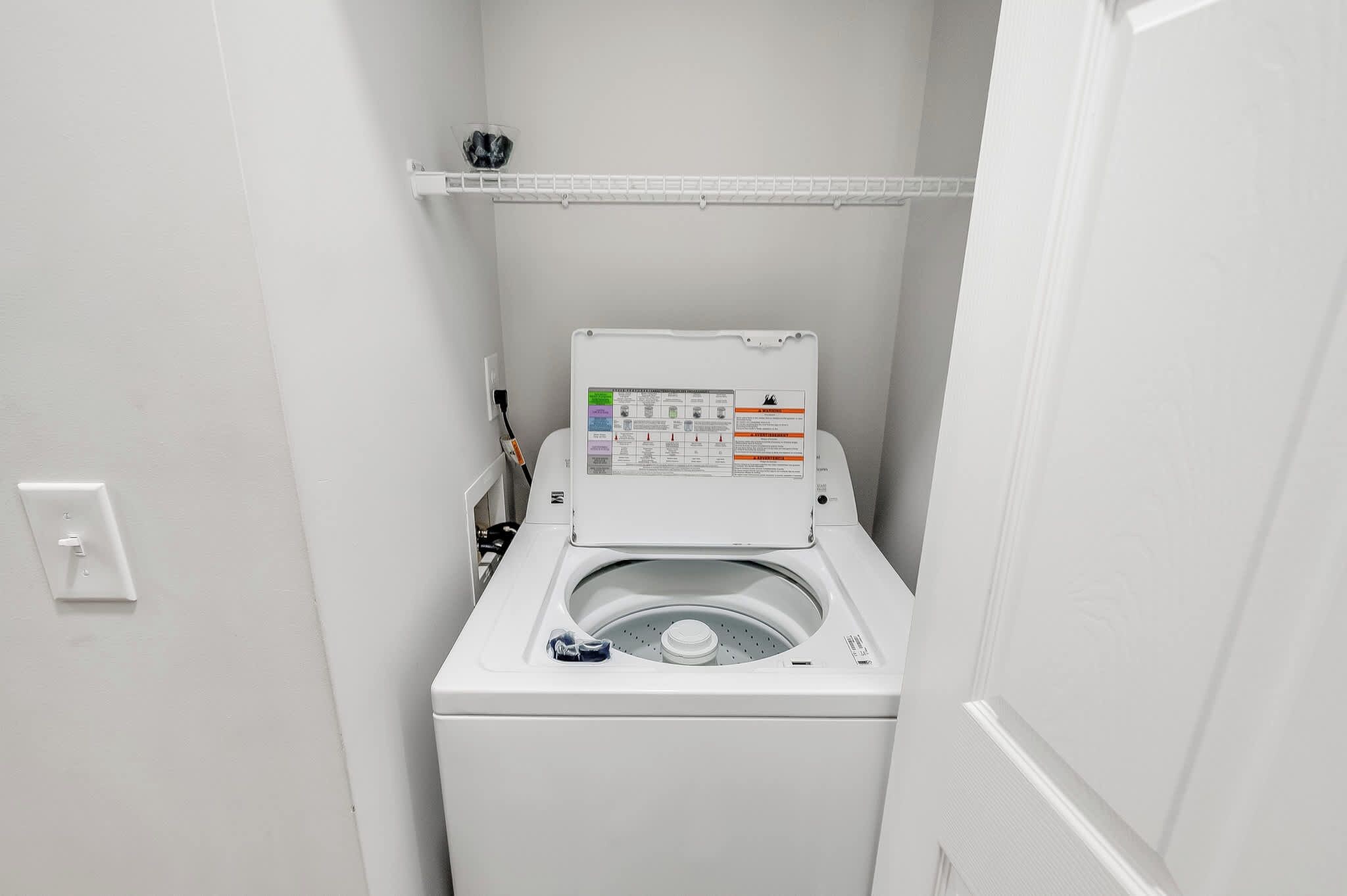 A compact, white, front-loading washing machine in a small laundry room with a control panel and a shelf above.
