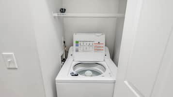 A compact, white, front-loading washing machine in a small laundry room with a control panel and a shelf above.