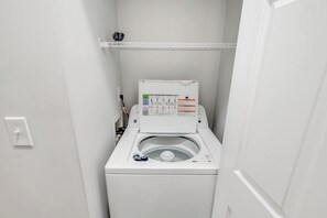 A compact, white, front-loading washing machine in a small laundry room with a control panel and a shelf above. - Large 2BR Unit with Pool Access, River Oaks Golf (Myrtle Beach)
