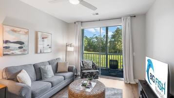A modern living room with a grey sofa, a coffee table, and a television. There is a balcony with a view of greenery.
