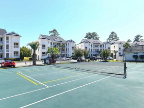Een tennisbaan met een net, omringd door een hek, naast een meer verdiepingen tellend woongebouw.
