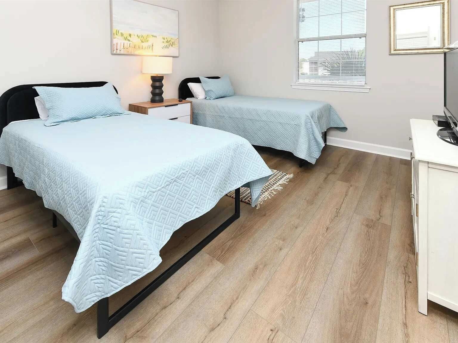 A bedroom with two beds, a nightstand, a lamp, a desk, and a window.