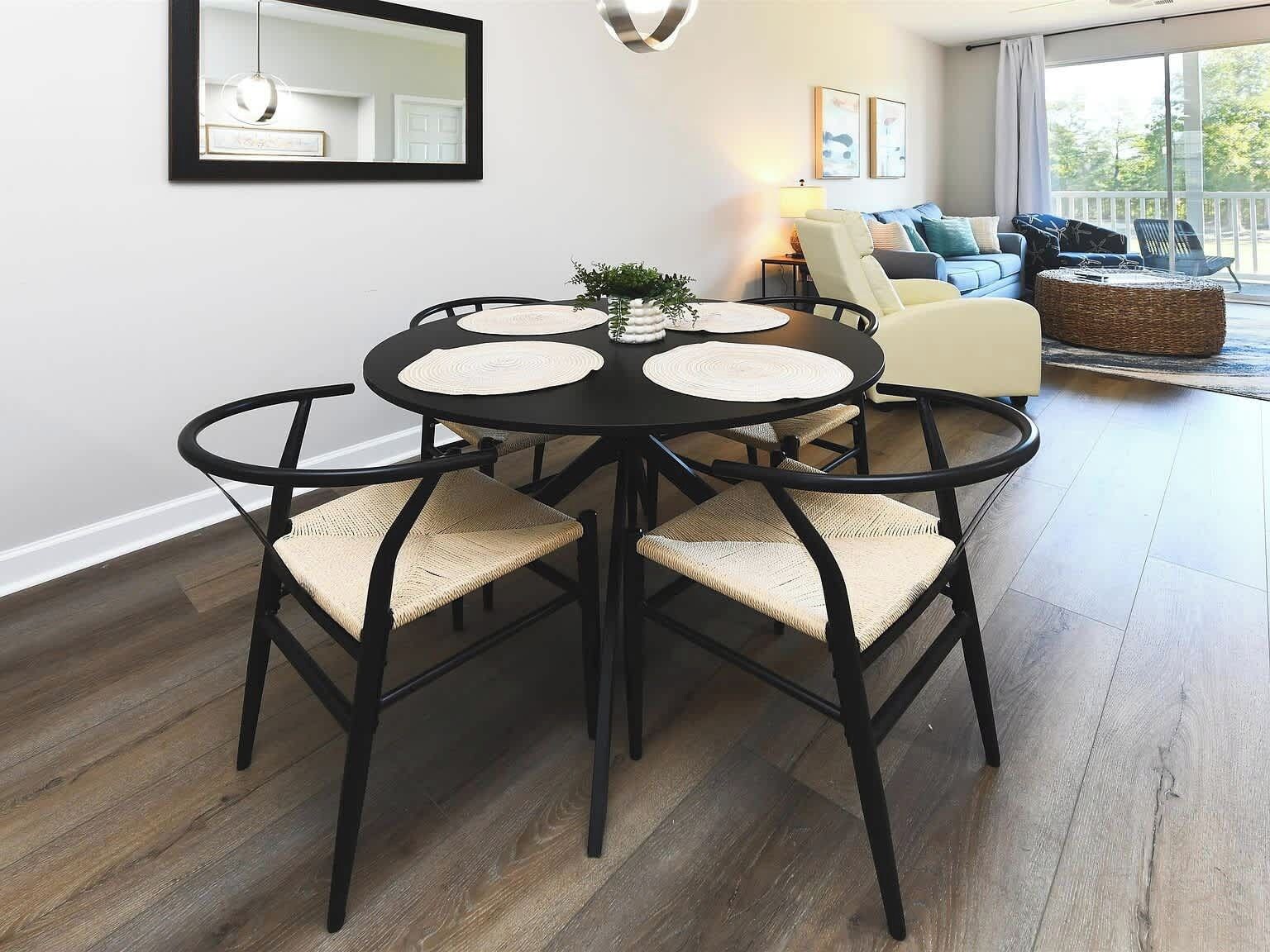 A dining area with a round table and four chairs, a mirror on the wall, and a sofa in the background.