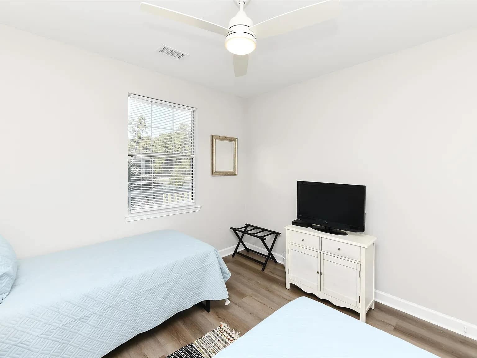 Un dormitorio con una cama, un televisor sobre un mueble blanco, un ventilador de techo y una ventana con persianas.