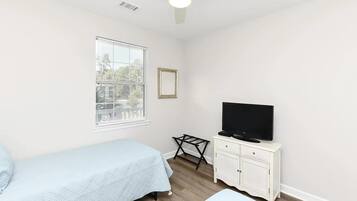 Un dormitorio con una cama, un televisor sobre un mueble blanco, un ventilador de techo y una ventana con persianas.