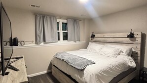 Een slaapkamer met een bed, een nachtkastje, een bureau met een computer, een raam met gordijnen en een plafondlamp.
