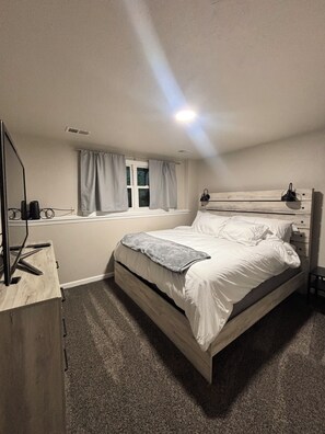 Quarto com cama, mesa de cabeceira, escrivaninha com computador, janela com cortinas e luminária no teto.