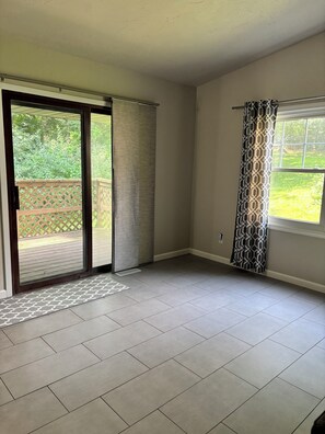 A room with a sliding glass door leading to a patio, a patterned curtain on a window, and a tiled floor.