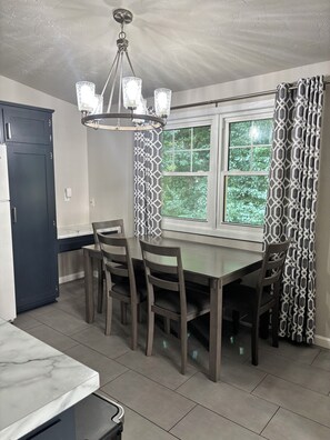 Un espace repas comprenant une table, des chaises, un lustre, une fenêtre avec des rideaux et une porte.