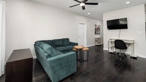 A modern living room with a sectional sofa, a ceiling fan, a TV mounted on the wall, and a small wooden table. - The A Lux Backyard House | 3BR w Private Backyard (Brownsville)