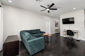 A modern living room with a sectional sofa, a ceiling fan, a TV mounted on the wall, and a small wooden table. - The A Lux Backyard House | 3BR w Private Backyard (Brownsville)