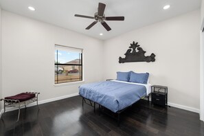 Quarto com cama, mesa de cabeceira, janela e ventoinha no teto, além de um objeto decorativo na parede.