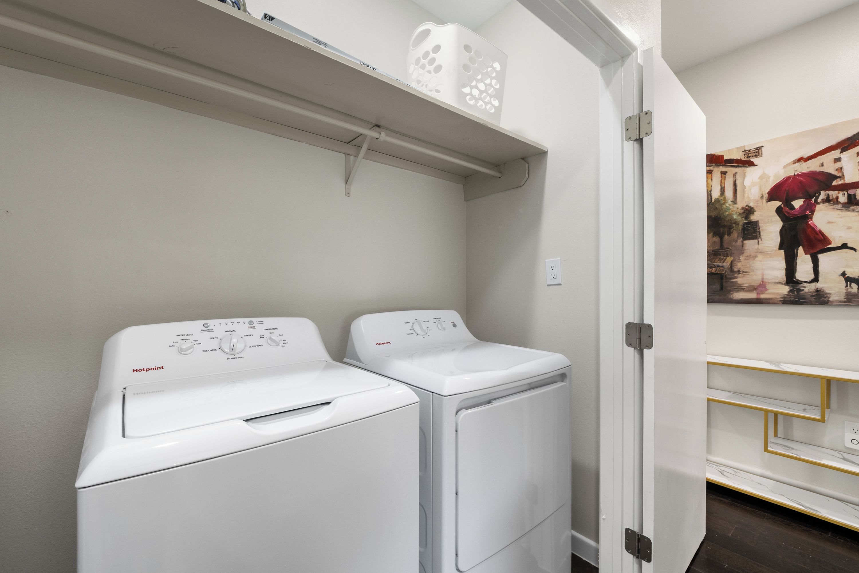 Un lavadero con dos lavadoras blancas de carga frontal, una pared blanca y una puerta que da a otra habitación.