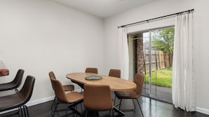 A modern dining area with a round table and six chairs, a large window with curtains, and a view of a backyard. - The A Lux Backyard House | 3BR w Private Backyard (Brownsville)