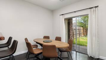 A modern dining area with a round table and six chairs, a large window with curtains, and a view of a backyard.