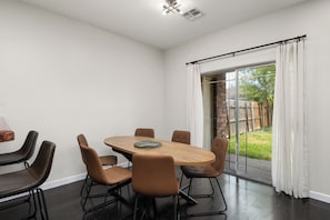 A modern dining area with a round table and six chairs, a large window with curtains, and a view of a backyard. - The A Lux Backyard House | 3BR w Private Backyard (Brownsville)