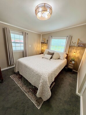 A bedroom with a large bed, a rug, a nightstand with a lamp, and a window with curtains. - The Pungo Perch: Waterfront Retreat with Boat Lift, Fishing & Pets Welcome (Belhaven)
