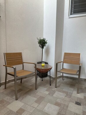 Zwei Stühle mit Holzlehnen und Metallgestellen, ein kleiner Tisch mit einem gewebten Sitz und ein Blumentopf mit einer gelben Blüte.